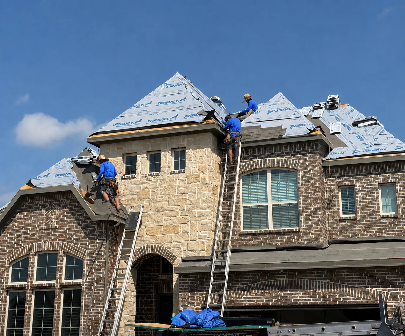 Two TopLine Roofing team members in branded shirts working across fresh underlayment on a stone fronted home, ladders still set against the wall